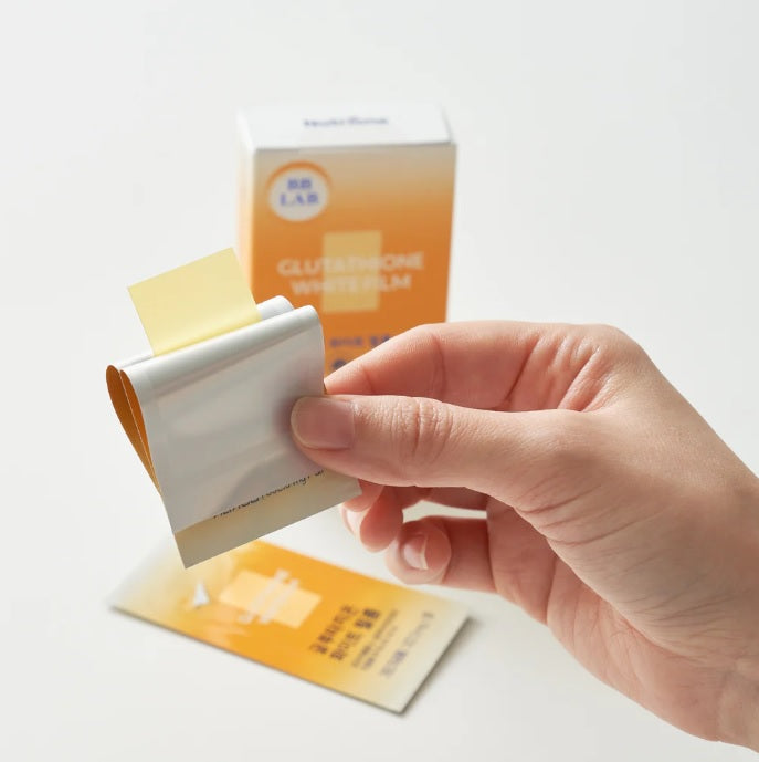 Пальцями тримають відкритий пакетик, з якого видно жовту плівку, на фоні стоїть коробка BB LAB Glutathione White Film.