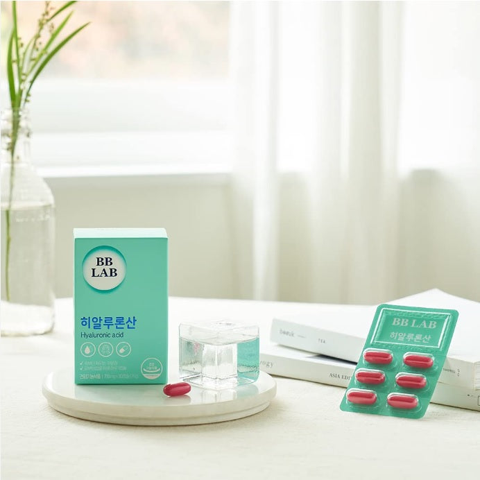 На білому подіумі стоїть коробка з колагеном BB Lab Hyaluronic Acid та червона капсула, поряд книжки та пластинка колагену.