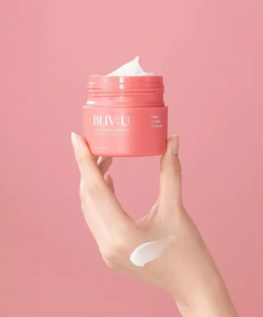 Пальці руки тримають відкриту баночку BLIV:U Collagen Bouncing Firming Cream 80 мл на рожевому фоні, на шкірі руки видно трохи білого крему
