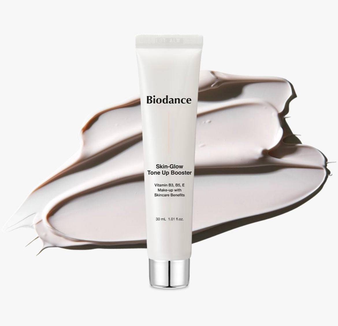Білий тюбик Biodance Skin Glow Tone Up Booster 30 мл з чорними написами та сріблястою кришечкою, стоїть, на фоні - кремова текстура нанесена на білу поверхню