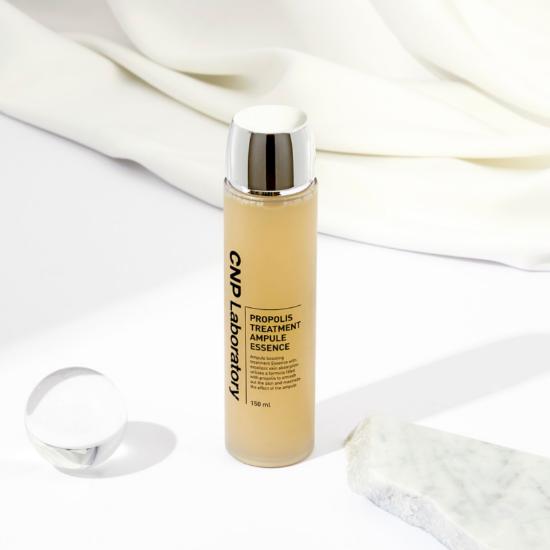 Пляшка CNP Laboratory Propolis Treatment Ampoule Essence стоїть на білій поверхні поряд з каменем та кулькою з рідиною, на фоні видно тканину.