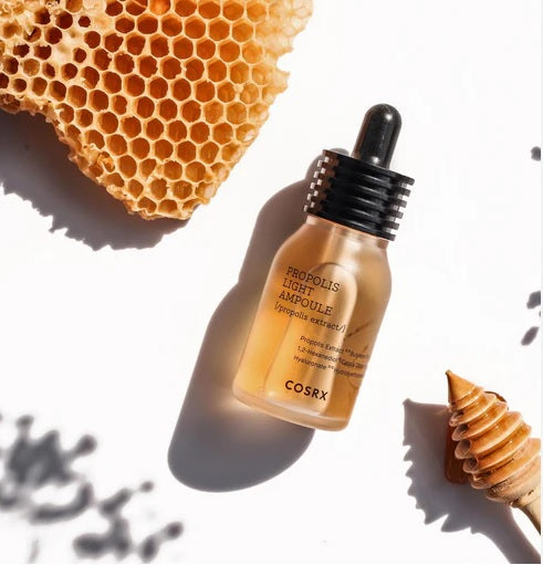 Пляшечка COSRX Propolis Essence лежить на білій поверхні поряд з шматочком медових сот і спеціальною ложкою для меду.