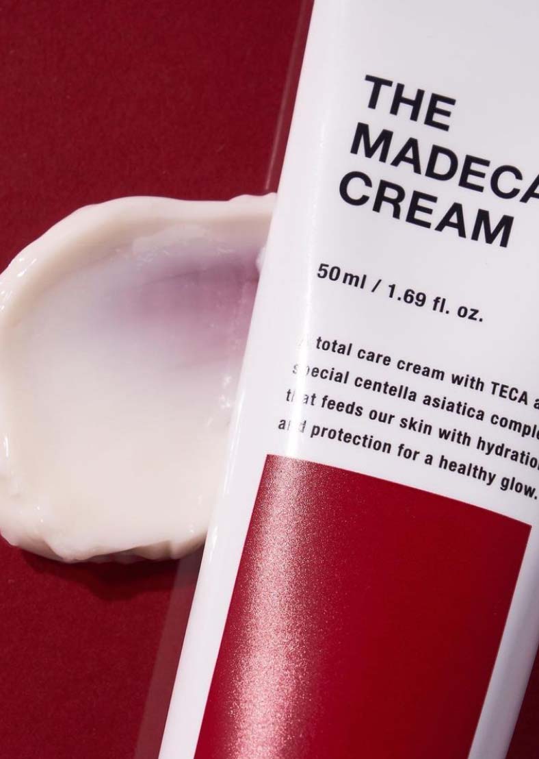 Крупний план тюбика крему Centellian24 The Madeca Cream 50 мл на червоному фоні, поруч — мазок білого крему на поверхні