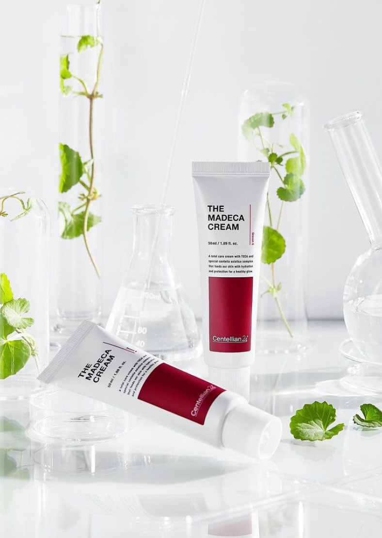 Два тюбики крему Centellian24 The Madeca Cream: один стоїть, інший лежить на поверхні. Поряд розміщені лабораторні пробірки та колби із зеленою рослиною всередині