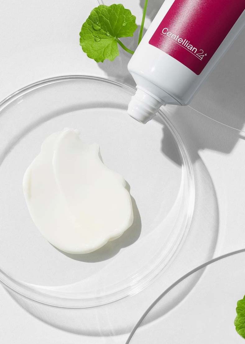 Відкритий тюбик крему Centellian24 The Madeca Cream лежить над прозорою скляною тарілочкою, на якій розподілено трохи білого крему, поряд видно зелену рослину