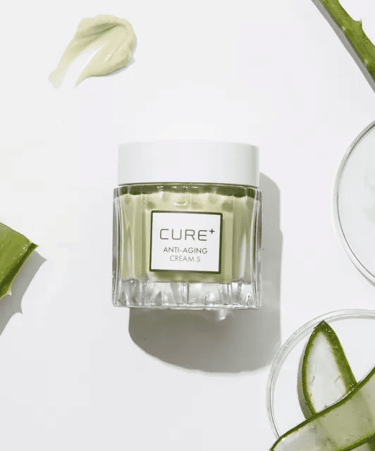 Баночка крему Cure Anti-Aging Cream лежить на білій поверхні поряд з розмазаним кремом. Біля неї розташоване скляне блюдце, на якому лежить нарізане алое.